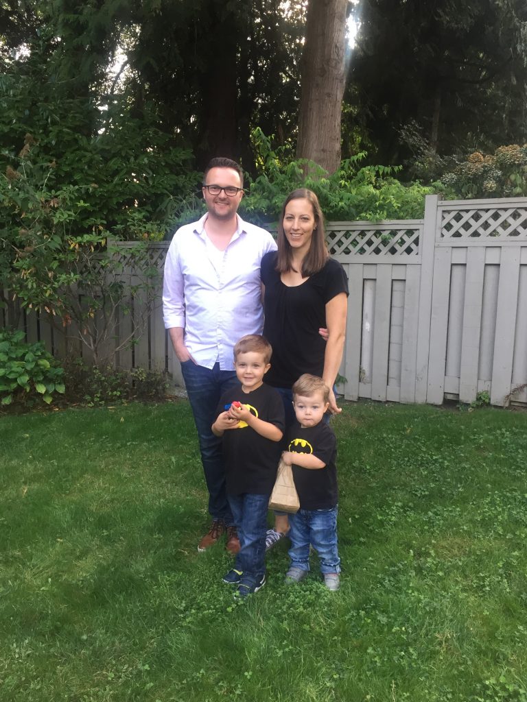 Family of 2 adults and 2 young boys standing in front of trees in their backyard